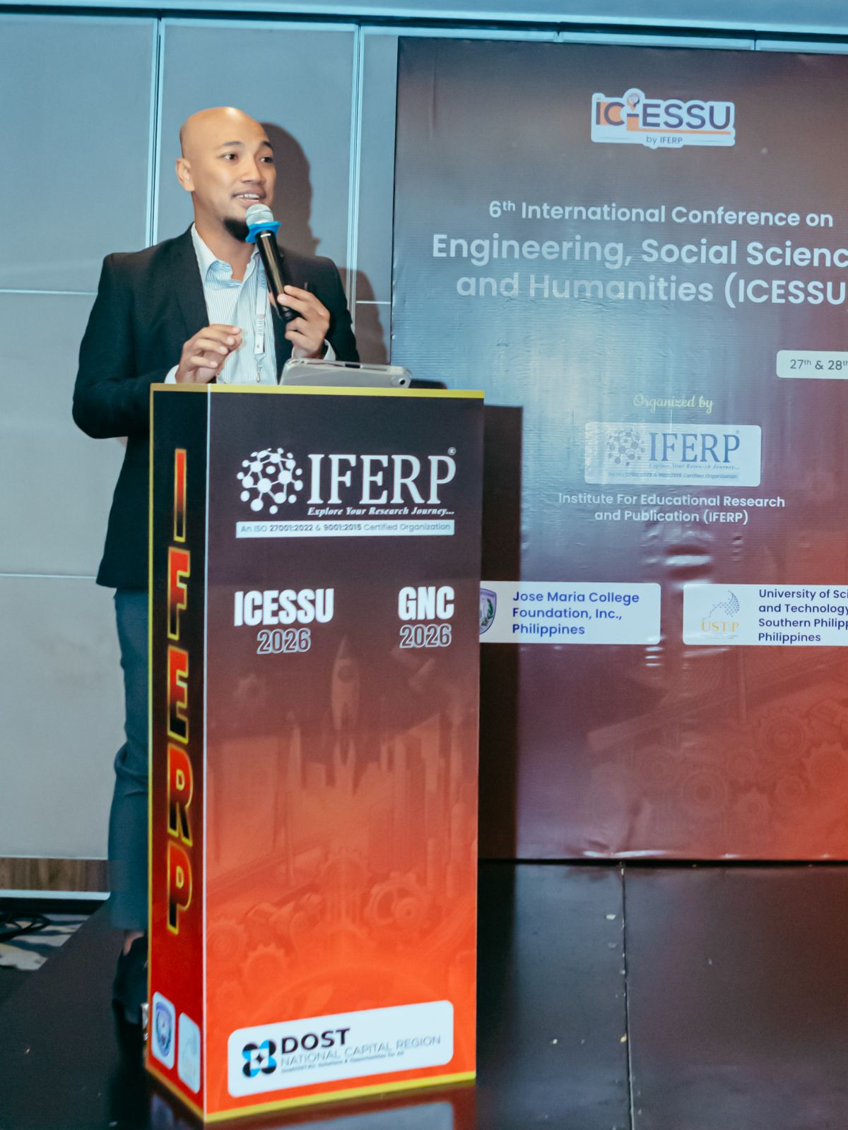 A presenter speaking at the IFERP podium in front of the 6th ICESSU backdrop